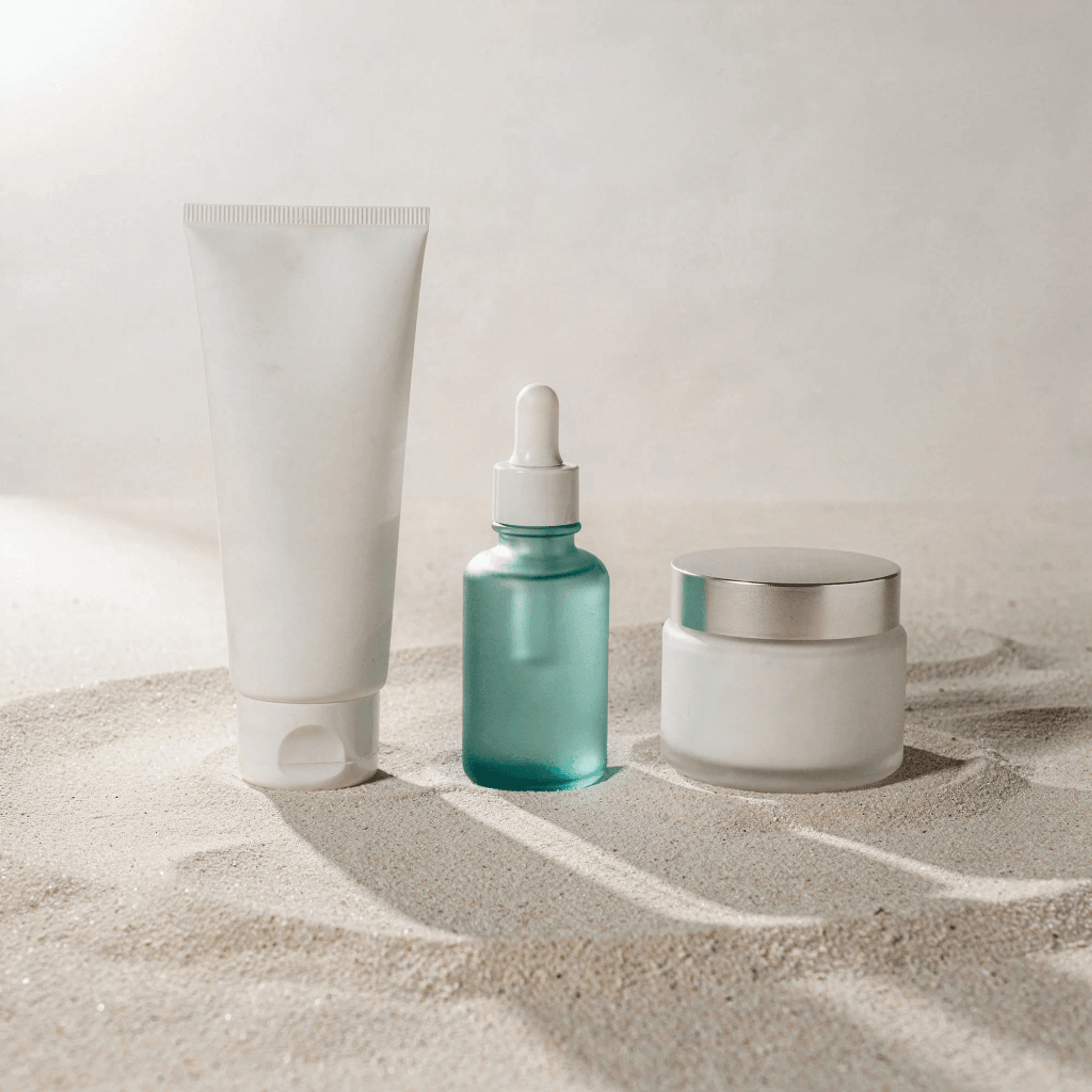 Concept rendering of three unbranded skincare containers — a tube, a sea-glass dropper bottle, and a frosted jar — standing in pale sand.